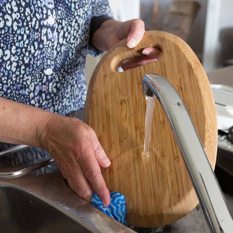 A person washing a round wooden cutting board in a sink with running water from a faucet, using a blue cloth.