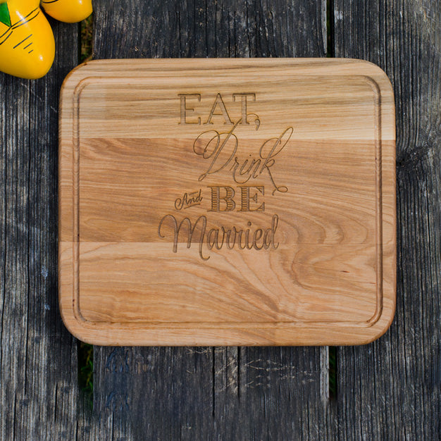 A rectangular wooden cutting board with rounded corners, featuring the engraved text 'EAT, Drink and BE Married' in a warm medium-brown wood tone. The board is photographed from above on a dark weathered wood plank surface, with a small yellow decorative object partially visible in the top-left corner.