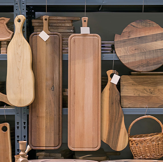 A collection of wooden serving boards and cutting boards in various shapes and natural wood tones, hanging on a dark metal shelf display.
